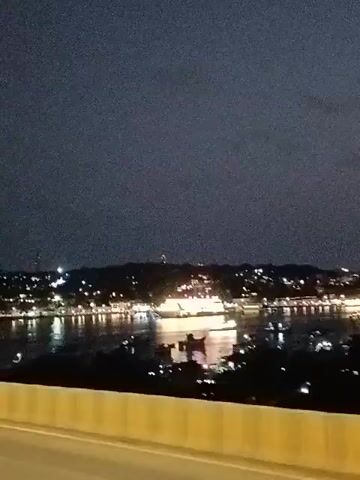 The video shows a nighttime view of a body of water from a bridge or elevated roadway. The scene is dark, with the sky a deep, cloud-covered twilight. Across the water, a cityscape or shoreline is illuminated with numerous lights, which reflect on the surface of the water. The camera is likely mounted in a moving vehicle, as indicated by the blurred movement. The foreground shows a concrete barrier or guardrail with yellow accents, typical of road infrastructure. The wind noise is significant, suggesting an exposed outdoor environment.