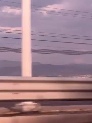 The video is taken from a moving vehicle, presumably a car, traveling along a highway or bridge. The camera faces towards a distant city skyline, appearing hazy and partially obscured by the light purple sky. Power lines run across the top of the frame, and a metal guardrail is visible in the foreground, indicating the perspective is from inside a vehicle. The environment appears to be arid or semi-arid, with dry vegetation on the hillside leading down to the city. The overall impression is one of transit and distance.