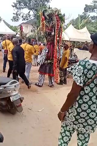 The video shows a crowd of people gathered around a performer in a traditional costume. The performer's outfit is elaborate, with a red and white striped pattern, feathered accents, and what appears to be greenery atop a tall headdress. Many people in the crowd are wearing yellow shirts. There are also two tents, a white building, and a couple of mopeds parked nearby. The scene takes place on a sandy or dirt road, with trees visible in the background.