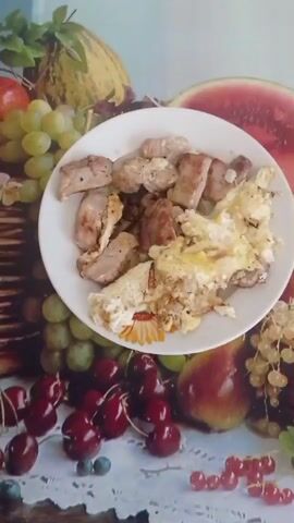 A plate with scrambled eggs and fried meat sits on a surface patterned with fruits like grapes, cherries, a pear, and a watermelon. The food is in focus, filling most of the frame. The plate is white and circular.