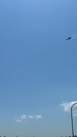 The video shows a fighter jet flying in a clear blue sky, leaving contrails behind. Below the plane, there is a body of water with several piers extending into it. People can be seen standing on the piers. In the distance, a city skyline is visible.
