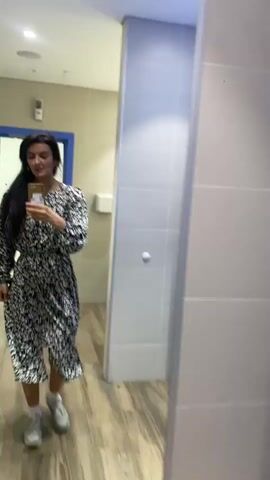 A woman walks quickly in front of a mirror on a bathroom wall, filming herself on a phone. She stops in front of the mirror and poses. The walls of the bathroom are covered with rectangular white tiles. A section of the wall is covered with hexagon-shaped tiles in black, light blue, and grey. The floor is covered with light-colored wood planks. The bathroom door is visible in the background, painted blue.