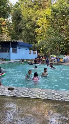 A group of children and adults are enjoying a swim at an outdoor swimming pool surrounded by lush green trees and vegetation. The pool is rectangular and filled with clear blue water. Some individuals are actively swimming and splashing, while others are standing or sitting at the edge of the pool. A light blue building with a white roof is visible in the background, situated behind the pool. The scene appears to be a recreational setting, possibly a public pool or a private resort, where people are engaging in water activities. The overall atmosphere seems relaxed and enjoyable.