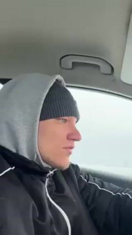 A young white male wearing a gray hooded sweatshirt over a gray beanie and a black jacket with white trim is driving a car. His left hand is visible on the steering wheel. The weather outside is snowy and overcast. Buildings are visible in the distance. Music is playing.