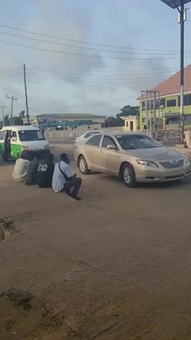 The video shows a gold Toyota Camry parked on a dirt shoulder beside a paved road.  Several people are squatting or sitting nearby. A green and white van is pulling onto the road. A solar-powered street light is next to the car. There is a two-story building with a reddish-brown roof visible in the background. Other cars, vans, and a three-wheeled vehicle are also visible. The scene appears to be an urban or semi-urban setting in daylight. There are overhead power lines running through the frame.
