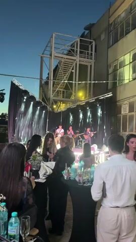 A live band performs on a stage outdoors at dusk. The band consists of at least four members: two guitarists, a bassist, and a vocalist. The stage is set up with black curtains as a backdrop and is lit by spotlights and decorative lighting elements. The audience is visible in the foreground, standing and sitting at tables with drinks. The overall atmosphere appears to be a concert or a social gathering with live music.