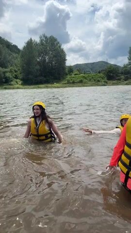 A group of people, some wearing yellow life jackets and helmets, are standing in a body of water. In the foreground, two individuals are visible. The person on the left is facing the camera and smiling, while the person on the right has their back to the camera, wearing a pink long-sleeved shirt and a yellow life jacket. In the mid-ground, a larger group of people are wading in the water. Some of them are interacting with each other. Further back, more people are visible, and several inflatable rafts, one red and two yellow, are beached on the shore. The background shows a lush green hillside with trees and some buildings, under a cloudy sky. The water appears murky and calm. The overall scene suggests an outdoor recreational activity, likely white-water rafting or a similar water sport.