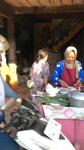 A woman wearing a hijab and an apron is preparing food at a stall. A person on a motorcycle is visible in the foreground. Another woman is standing behind the food stall. The scene appears to be outdoors, possibly a market or street vendor setting, with natural lighting. The woman in the hijab is holding a utensil and appears to be working with food items on a surface. There are various containers and food wrappers present on the stall. The background shows a building with an indoor staircase.