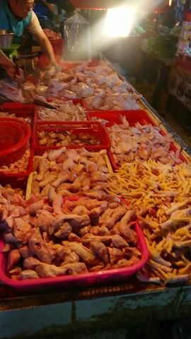 The video captures a lively night market scene. The initial frames focus on a meat stall, predominantly displaying various raw chicken parts, such as necks, feet, and bones, arranged in red and pink plastic crates. A white tray also holds cuts of meat. Two individuals are seen interacting at this stall, one with an outstretched arm, suggesting a transaction.

As the camera pans and moves, it reveals other market stalls featuring an abundance of fresh produce. Piles of vegetables like onions, leafy greens, potatoes, and what appear to be tomatoes and chilies are neatly displayed on wooden counters. Several people are visible throughout the market, browsing the stalls, walking through the aisles, and engaging in shopping activities.

The setting is an open-air or covered market, brightly illuminated by artificial lights, indicating the time of day is evening or night. The overall atmosphere appears busy and bustling with activity. No discernible text is visible in the video.