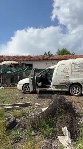 The video shows a white van parked in a yard on a sunny day with some clouds. The driver's side door of the van is open, and two white chickens are inside the vehicle. One chicken is standing on the floor of the van, near the open door, while the other is on the driver's seat. The interior of the van appears dusty and worn. In the background, there's a building with a red tile roof and a green tarp partially covering something. The yard is overgrown with weeds and littered with various items, including a tire and a coil of wire.