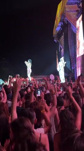 The video shows a nighttime outdoor concert with a crowd of people visible in the foreground. The crowd is facing a stage where two performers are visible. The performer on the left is a woman wearing a light-colored crop top and a pink skirt, with her hair pulled back in pigtails. She is holding a microphone and appears to be singing. The performer on the right is also dressed in light-colored clothing and is holding a guitar, suggesting they are a musician. Both performers are illuminated by stage lights. Many people in the crowd are holding up their cell phones, presumably to record or photograph the performance. Some are also raising their hands in the air, indicating excitement. The stage itself is large and has a screen or banner visible at the top with the word "VIVO" on it. The overall atmosphere suggests a lively and energetic concert event.