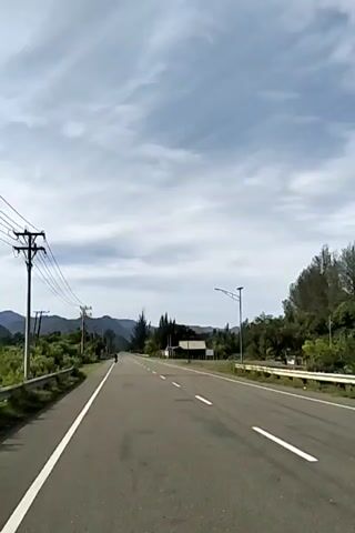 The video shows a straight, paved road stretching into the distance, with mountains visible on the horizon.  On either side of the road is vegetation and trees. Power lines run along the left side of the road, supported by utility poles. A small building or structure is visible on the right side of the road, along with a street light. The sky is cloudy.  A vehicle, possibly a motorcycle, can be seen far down the road.