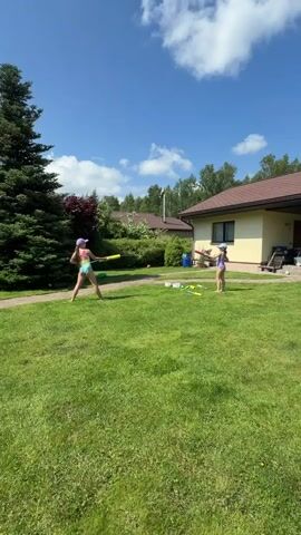 Two young girls are playing baseball in a grassy backyard on a sunny day. The girl on the left is wearing a swimsuit and a baseball cap, and is swinging a yellow bat. The girl on the right is wearing a purple swimsuit and is holding a yellow bat. There are trees and houses in the background.