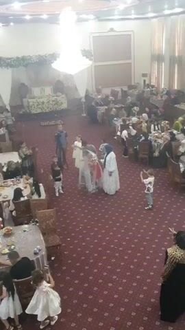 The video captures a wedding ceremony. In the center, a bride, adorned in a traditional white wedding dress and veil, walks down the aisle. She is accompanied by two women, one on each side, appearing to be her attendants or family members. Several children, dressed in formal attire, are also part of the procession. A person holding a video camera documents the event. The ceremony is taking place in a large hall with red carpet flooring. Tables are set up throughout the venue, suggesting a reception will follow. Guests are seated at the tables, observing the procession. The overall atmosphere is festive.