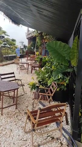 The video shows an outdoor seating area at what appears to be a cafe or restaurant. The ground is covered in white gravel. There are several tables and chairs made of dark wood and metal, arranged on the gravel. To the right of the frame, there are many potted plants, some of which are quite large and leafy. A person wearing a blue jacket and a hat is standing in the background on the left, near a railing. The area is partially covered by a dark canopy, possibly for shade. The overall impression is that of a somewhat rustic and nature-inspired outdoor space.