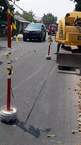The video shows a road with traffic cones and caution tape indicating a construction zone. A yellow excavator is parked on the right side of the road, and a red truck is driving away from the camera in the distance. The road appears to be under repair, with some digging or leveling work happening. The sky is visible and appears to be clear. The video is taken from a slightly elevated perspective, looking down at the road.