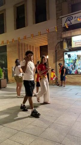 The video captures a street scene at dusk. In the foreground, a young man with dark, curly hair, wearing a white t-shirt and black shorts, walks towards the right of the frame, holding his phone to his ear. Behind him, a young woman with long brown hair, wearing a red and yellow shirt and cream-colored pants, also walks to the right, looking at her phone. Several other people are visible in the background, including a young child wearing a red and yellow cape. The street is paved, and brick buildings line one side of the street. A storefront with a sign reading "Ferreterias" is visible. Decorative flags in red and yellow are strung across the street. The lighting suggests it is evening, with artificial lights illuminating the scene.