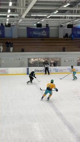 An indoor ice hockey rink is shown with a game in progress. Five players are visible on the ice, along with a referee. Two players in black and green jerseys are on one team, and three players in teal and yellow jerseys are on the other. The players are skating, holding hockey sticks, and engaged in gameplay. The referee, wearing black and white stripes, is also on the ice, observing the game. The rink has boards around the perimeter, with advertisements visible on the boards. Bleachers with spectators are in the background, and the ceiling has industrial lighting. The ice surface is marked with lines, and a puck is not clearly visible but implied by the players' actions. The overall environment appears to be a sports facility dedicated to ice hockey.