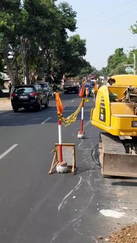 A street scene shows construction or road work in progress. A yellow excavator is parked on the right side of the road, with its tracks visible. Traffic cones and warning tape are set up to delineate a construction area, with orange flags attached to poles. Several vehicles, including cars, a truck, and a motorcycle, are on the road, some passing through the construction zone. Trees line the left side of the street, and buildings are visible in the background. The sky is clear and bright.