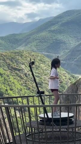 A young girl in a white shirt and red shorts stands on a rotating platform, facing left, with a scenic mountain backdrop. She looks around as the platform rotates her. Her hair is in two pigtails. A mountain range with green foliage stretches across the background. Two distant figures are visible on a rope bridge spanning between two mountains. The girl is on a balcony with a metal railing.