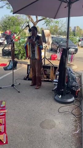 A man dressed in traditional Native American attire, complete with a headdress and fringed vest, plays a flute. He stands under a large umbrella, with his merchandise spread out on the ground in front of him. The merchandise includes several pan flutes and a collection of black t-shirts featuring Native American-themed designs, such as a dreamcatcher with wolves and a portrait of a Native American chief. In the background, cars and other vendors are visible, suggesting the setting is an outdoor market or fair. The man is surrounded by musical instruments and sound equipment, indicating he is a street performer. The overall atmosphere appears to be relaxed and festive, with the music adding to the ambiance.