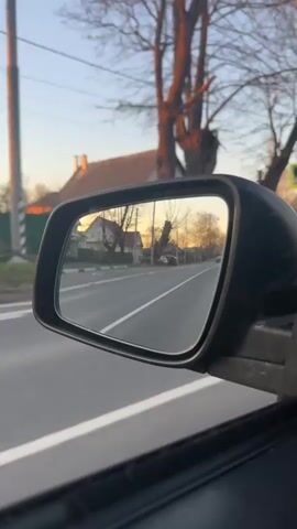 The video depicts a view from a car's side mirror as it drives down a road. The mirror reflects the road behind the car, showing a white line dividing the lanes, houses, trees without leaves, and street signs. The sky visible in the reflection is light blue with a hint of orange from the setting sun. The car is moving forward, as evidenced by the changing scenery in the mirror. The foreground shows the interior of the car with the window and the side mirror casing.
