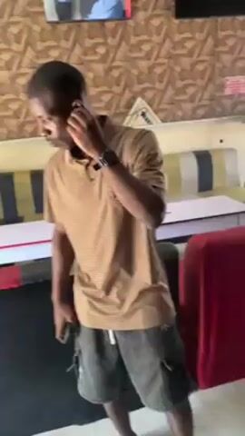 A slender man stands indoors. He has dark skin and a short haircut, wearing a light brown polo shirt, dark gray cargo shorts, and gray slides. He has a wristwatch on his left wrist and white earbuds in his ears. He holds a black smartphone in his right hand. The man is looking directly at the camera. The background shows a wall with a geometric pattern, a flat-screen TV displaying a news program, and seating areas with red and striped cushions.