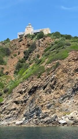 A lighthouse sits atop a rocky hill overlooking the ocean. The lighthouse is white with a glass lantern room. The hill is covered in sparse vegetation, with some green shrubs and dry grass. The ocean is a deep blue, and the sky is clear. The video is shot from a boat, providing a view of the lighthouse from the water. The camera pans slightly, showing the scale of the hill and the lighthouse.