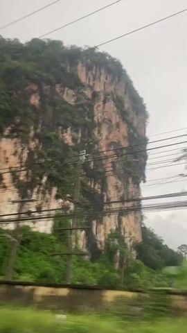 A passenger riding in a vehicle records a large, steep cliff formation. The cliff face is streaked with shades of beige, light brown, and green vegetation. Power lines stretch in front of the cliff. The vehicle passes by a road with colorful barriers, roadside businesses, and a truck. The sky is overcast.