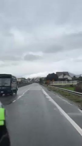 The video shows a view from a vehicle driving on a wet road under a cloudy, overcast sky. The road is paved and has a white line marking the edge.  A green truck is visible ahead on the left side of the road. As the vehicle moves forward, buildings and houses appear on the right side of the road. Some of the buildings have visible signs. There are a few trees and some greenery visible. The road appears to be relatively clear of other traffic.