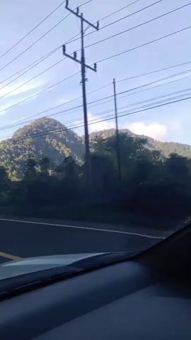 This video shows a view from inside a vehicle looking out at a mountainous landscape. The sun is shining, and there are power lines visible in the sky. The vehicle appears to be driving on a road that runs alongside the mountains. There are trees and some buildings visible near the road. The video is shot in daylight.