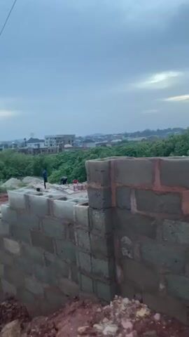 This media file captures a construction scene in Rayfield, Nigeria, under a cloudy sky, suggesting it's likely early morning or late afternoon. The foreground is dominated by a partially built brick wall, with exposed soil and some debris at its base. 

In the background, several individuals are engaged in construction work. Their activities appear to involve laying bricks and possibly mixing cement, indicative of ongoing building operations. A few structures, including what looks like a multi-story building and smaller houses, are visible in the distance, interspersed with lush green foliage. Power lines stretch across the upper portion of the frame, a common sight in many urban and semi-urban settings.

The overall mood conveyed is one of diligent labor and development, set against a somewhat overcast but still bright sky. The presence of multiple workers implies a collaborative effort on what is likely a new construction project. The environment appears to be an open area, possibly on the outskirts of a more developed town or city, given the mix of construction and natural greenery. No visible text is present in the media file.