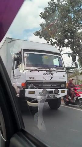 The video shows a white Mitsubishi Fuso truck in motion on a street in Klatakan, Indonesia. The truck has "RFX Top Stop" written on its side, along with "4X2 L" and "MITSUBISHI FUSO". The license plate reads "B 9433 KD".

In the background, a person on a motorcycle is visible. The scene appears to be during the daytime, with clear skies and sunlight. Trees line the street, and there are some buildings visible in the distance. The video is filmed from inside another vehicle, looking out through the window. The overall mood is casual and observational, capturing a slice of daily life on the road in Indonesia.