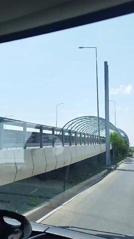 The video, captured from inside a vehicle, shows a road with a distinctive modern bridge or overpass on the left. The bridge has a unique arched structure covered with a reflective material, possibly a walkway or a tunnel. Trees and greenery line the area beneath and around the bridge. The road is paved and appears to be a multi-lane highway or a major road, with clear lane markings. Traffic lights and street lamps are visible along the roadside. Several vehicles, including cars, are seen on the road. The sky is clear and blue, indicating a sunny day. The video shows the vehicle moving forward along the road, with the bridge gradually receding from view. There are no people visible in the video, and the focus is on the infrastructure and the surrounding environment.