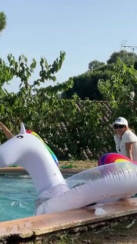 A young person with dark hair is in a swimming pool, partially submerged and struggling with a large, inflatable white unicorn pool float. The person's head is down, and their body is positioned awkwardly under the float. Another person wearing a white hat and sunglasses is leaning over the edge of the pool, seemingly observing or assisting. The pool is surrounded by a concrete edge, and lush green trees and foliage are visible in the background under a clear blue sky.