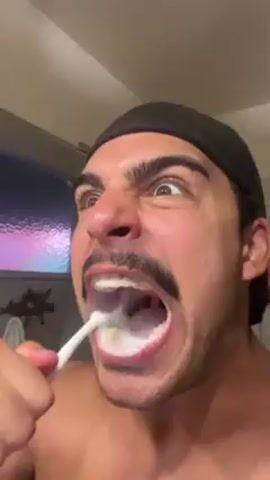 A man with a mustache and a black head covering is brushing his teeth with a white toothbrush. He appears to be looking directly into the camera with wide eyes. His mouth is open, revealing foamy toothpaste. The bathroom background includes a window with a lavender tint, a towel, and a starfish decoration.