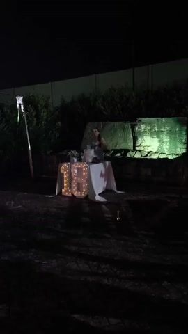 A young woman stands behind a table set up outdoors at night. On the table is a cake, a box, and some decorations. In front of the table, illuminated by lights, are two large numbers, "18." The woman appears to be holding something in her hands. Behind the table, a wall is lit with green light, and there is dense foliage. The ground in front of the table is dark and appears to be gravel or dirt.