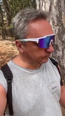 The video shows a man with graying hair wearing sunglasses and a gray t-shirt with a backpack on. He is walking in a wooded area with pine trees. The ground is covered with dirt and rocks. The man appears to be recording himself with a camera mounted on a selfie stick. He is looking down at the ground as he walks.