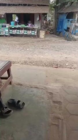A video begins with a person sitting on a wooden bench. They are wearing a black headscarf and black clothing. Next to them are a pair of black sandals. In the background, a small shop or stall is visible with several people and various items on display. The ground in front of the bench is concrete, and behind it is a dirt path. The video then pans to the right, showing a person riding a motorcycle. Another motorcycle is parked nearby. The scene appears to be an outdoor environment, possibly a rural area or a village.