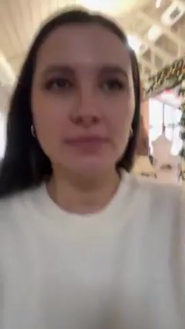 A woman with long dark hair, wearing a white sweater and hoop earrings, is facing the camera. She touches her face and hair. The background appears to be an interior space decorated for the holidays, possibly a store or public area, with Christmas-themed decorations and lights visible. The woman smiles slightly at the camera.
