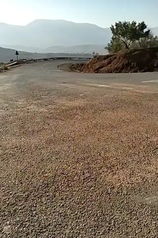 The video shows a motorcyclist riding on a winding road, away from the camera. The road is paved, with a gravel shoulder in the foreground. The motorcycle is black with a brown leather seat. The rider is wearing a helmet and an orange shirt. In the background, there are mountains and a clear blue sky. There is a small tree and dirt embankment on the right side of the frame. There are road signs visible on the side of the road.