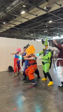 The video shows a group of people in costumes of characters from the anime Dragon Ball Z, posing for a photo. In the foreground, a person dressed as Goku is in a fighting stance, with their hands outstretched and glowing blue orbs of energy between them. To their right, a person dressed as Cell, a green insect-like villain, is also posing with outstretched hands, and blue energy orbs are visible between their hands and Goku's. Behind Goku, several other characters are visible: a person with pink hair, possibly Super Saiyan God Goku or Bulma; a person dressed as Vegeta, with spiky black hair and purple clothing; and another person in brown and white robes, possibly Master Roshi. The group appears to be at a convention or event, with a large open space and white partition walls in the background. The lighting suggests an indoor venue.