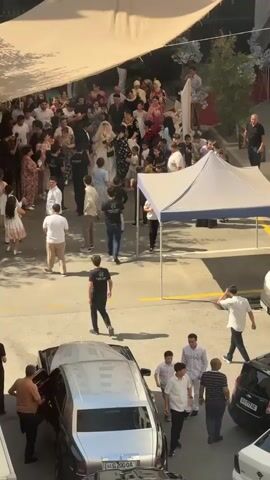 A wedding procession is moving under a large, cream-colored awning, possibly outdoors. A bride in a white wedding dress and veil is visible in the center, accompanied by a groom in a dark suit. They are surrounded by a crowd of people, many of whom appear to be guests, including men, women, and children dressed in various attire. Some individuals in black t-shirts with a logo are positioned close to the wedding couple, possibly acting as security or event staff. Several photographers and videographers are also present, capturing the event. In the foreground, there are parked cars, including a dark-colored luxury sedan. People are walking and interacting, suggesting a lively celebration. The environment appears to be a paved area, possibly a street or courtyard, with some greenery visible in the background. The overall scene depicts a joyful and public wedding ceremony.