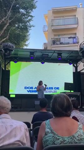 A woman stands on a stage in front of a large LED screen, singing into a microphone. The screen displays text that reads "DOS ANIMALES DE VERBENA SALVAJE DE ABRIL MORTAL". She is wearing a blue romper and appears to be performing outdoors in front of an audience. The stage is equipped with lighting equipment, and behind the stage, a building with balconies is visible. The audience members are seated, facing the stage, with their backs to the camera. The scene is illuminated by natural daylight.