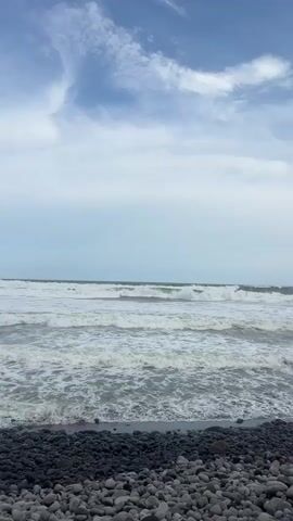The video shows a wide shot of a rocky beach with waves crashing on the shore. The sky is overcast, with some scattered clouds. The ocean is a deep blue-grey, and the waves are white-capped. The beach is made up of smooth, grey pebbles. The video is taken from a low angle, looking out at the ocean.