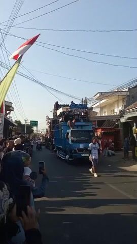 A group of people are gathered on the side of a street, some holding up phones to record a parade. The parade features a blue truck adorned with speakers and a large sound system. Several people are standing on top of the truck. A red and white flag is visible in the upper left corner of the frame, attached to a pole. The sky is clear and blue. The scene appears to be in a residential area with houses and shops lining the street.