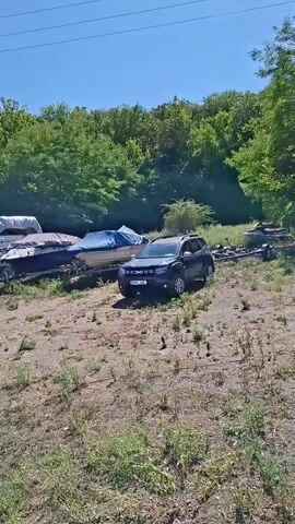 The video pans across a dry, grassy area with several boats on trailers parked in front of a dense line of trees under a clear blue sky. A dark colored SUV is parked to the right of the boats. A large electrical tower is visible in the background to the left.