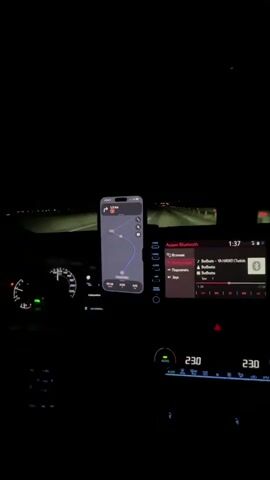 The video is filmed from inside a car at night. The dashboard is visible, with the car's navigation screen displaying a map and route. The car's speedometer is illuminated, showing a speed of around 70 km/h. The car's infotainment system is playing music, with song titles and artist names displayed. The time is 1:37. The climate control is set to 23.0 degrees Celsius. The car is driving on a road with streetlights and oncoming traffic. The driver's hands are not visible.