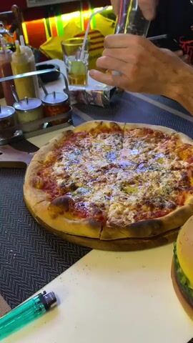 A close-up shot shows a person adding pepper to a freshly baked pizza with a golden crust. The pizza, cut into slices and served on a wooden board, features a melted cheese topping and a tomato sauce base. Condiments are arranged in small metal containers nearby. In the background, another person is visible, looking at their smartphone. There is also a drink and what appears to be a decorative burger on the table.