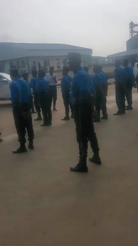 The video shows a group of people, most of whom appear to be male, dressed in blue shirts and dark pants, standing in a line outdoors. They are all wearing dark caps. The individuals are facing away from the camera, except for one person in the foreground who is facing sideways. The background shows buildings, including what appears to be an industrial facility with large metal structures, and several parked vehicles. The ground is paved with concrete or asphalt. The sky is overcast. There are no overt actions or interactions captured in the short clips.